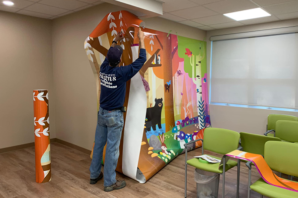 A person is installing a colorful forest-themed wall mural in a room with green chairs and a window. The wall murals feature trees, animals, and bright colors, with a rolled-up section leaning against the wall.