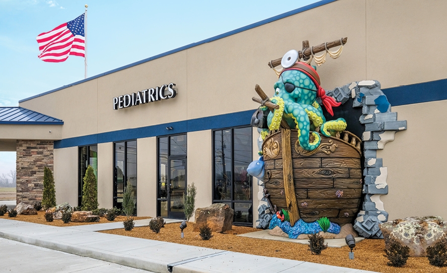 A pediatric medical clinic with an American flag and large windows features a colorful doctor octopus sculpture bursting through a wall in a wooden barrel, creating a playful landmark with children’s decor for a welcoming entrance.