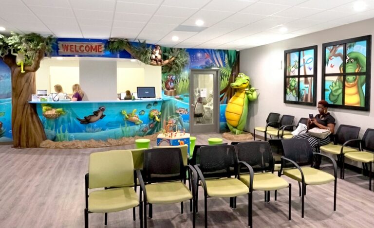 Twighlight everglade-themed waiting area with calming sunset bayou wall murals, sculpted kid-friendly alligator character and reception desk with sculpted tree and underwater characters.