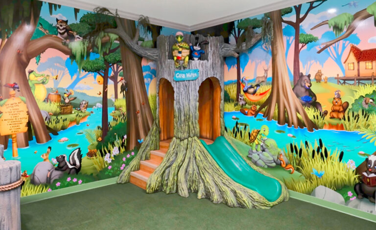 A colorful indoor playroom with painted murals of cartoon forest animals, trees, and a river brings a bayou-theme to medical clinic decor. A tree-shaped play structure with two slides sits among playful animal characters and vibrant scenery.