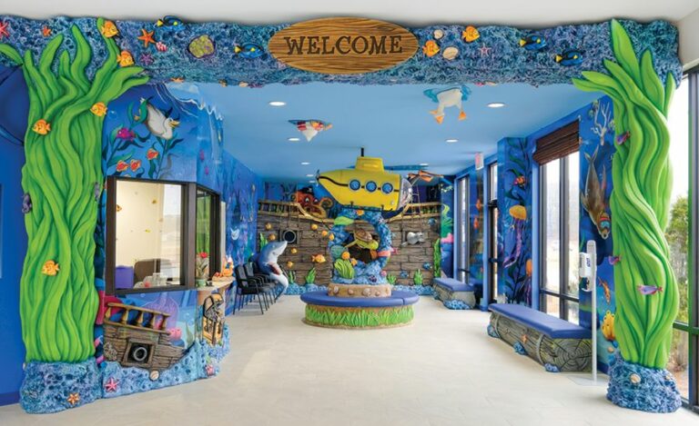 Entrance into an undersea, shipwreck-themed reception area with sculpted coral, seaweed and wood board 'welcome' sign. Themed wall murals, sculpted characters and custom centerpiece bench with shipwreck wall cladding add to the decor.