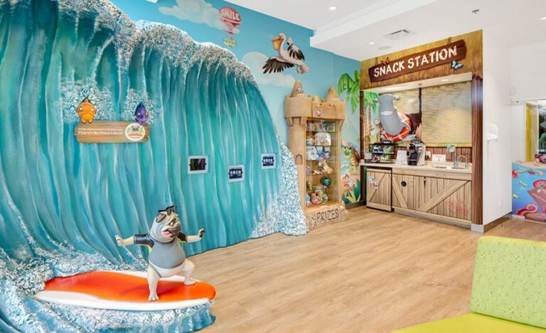 Beach-themed waiting area with sculpted wave, surfing pug dog and wall-mounted gaming pads, a sandcastle prize tower and surf-shack snack station.