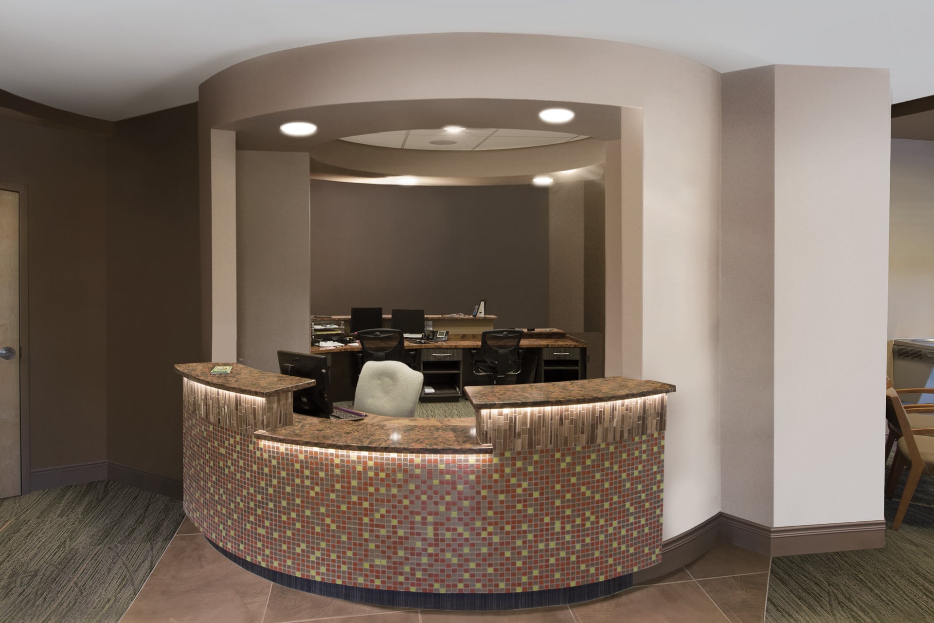 A modern reception desk with a curved counter featuring small, multicolored tiles brightens up what was once a drab area. Behind the desk are office chairs, computers, and workspaces set in a warmly lit space with neutral-colored walls.