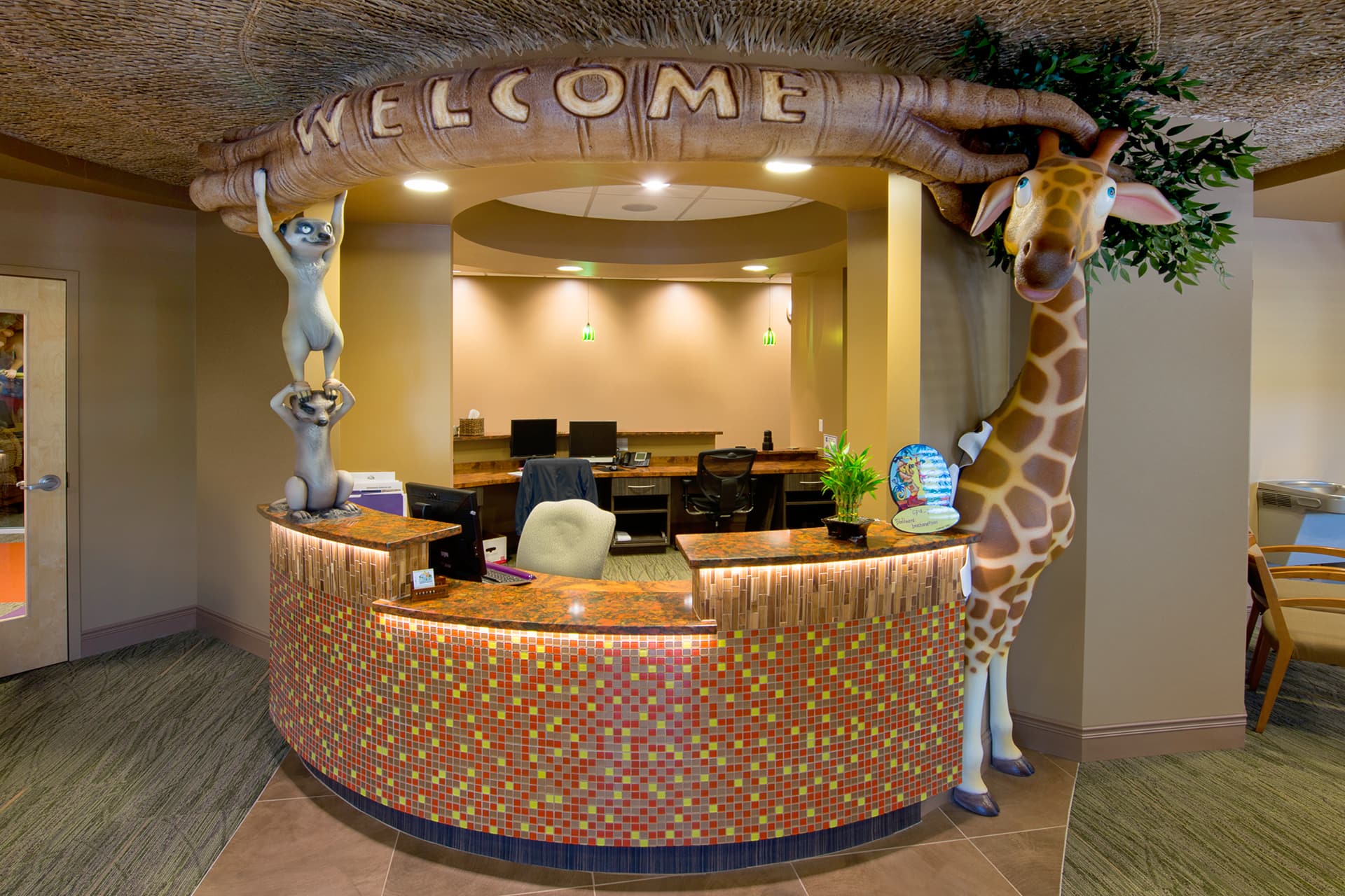 A colorful reception desk with a large “WELCOME” sign, playful animal statues like a giraffe and lemurs, and warm lighting showcase the transformation into a friendly, inviting kids space.