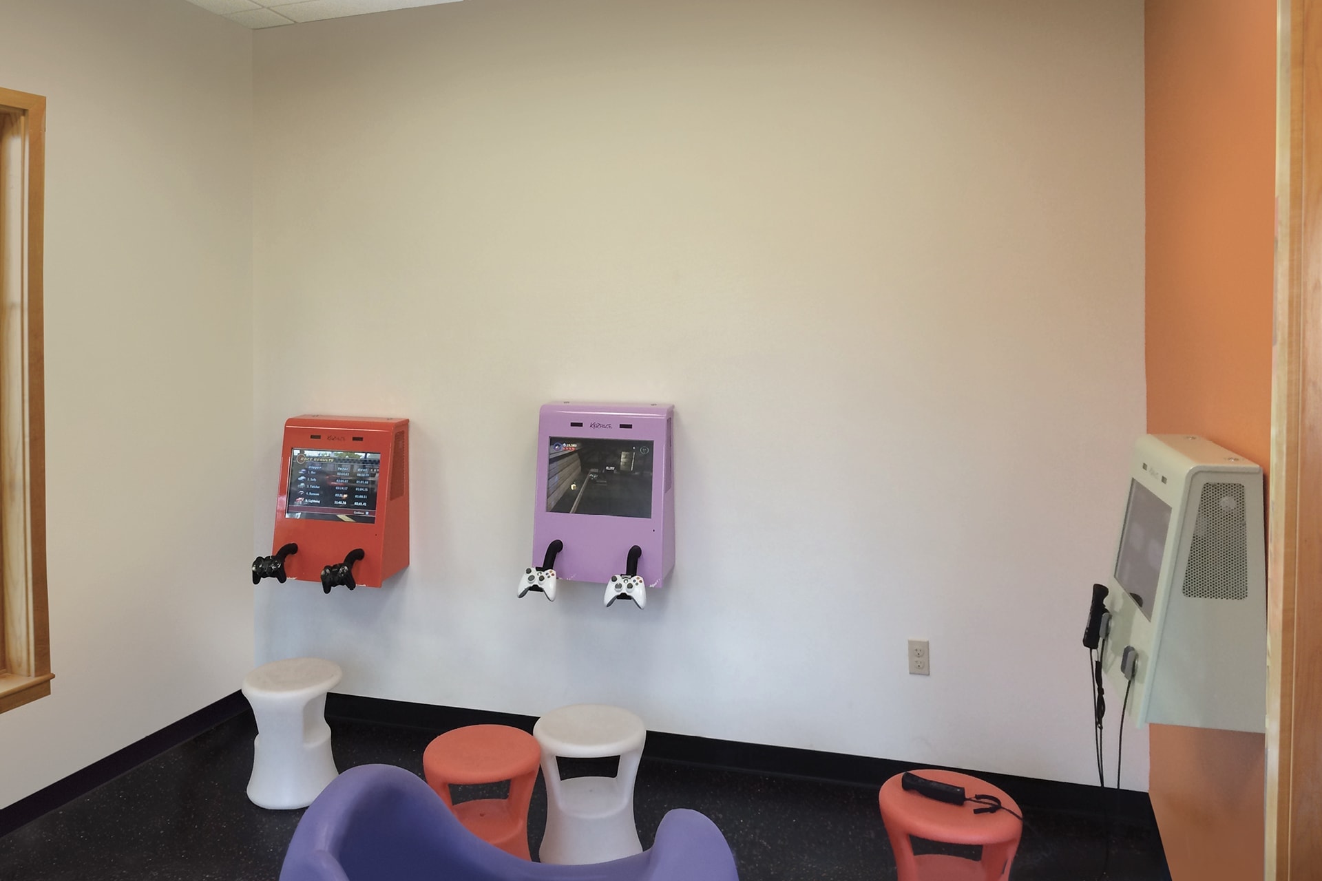 This boring before picture shows a small room with two wall-mounted gaming consoles, pastel seats, and a wall-mounted device with a telephone handset. The light-colored walls add to the drab atmosphere.
