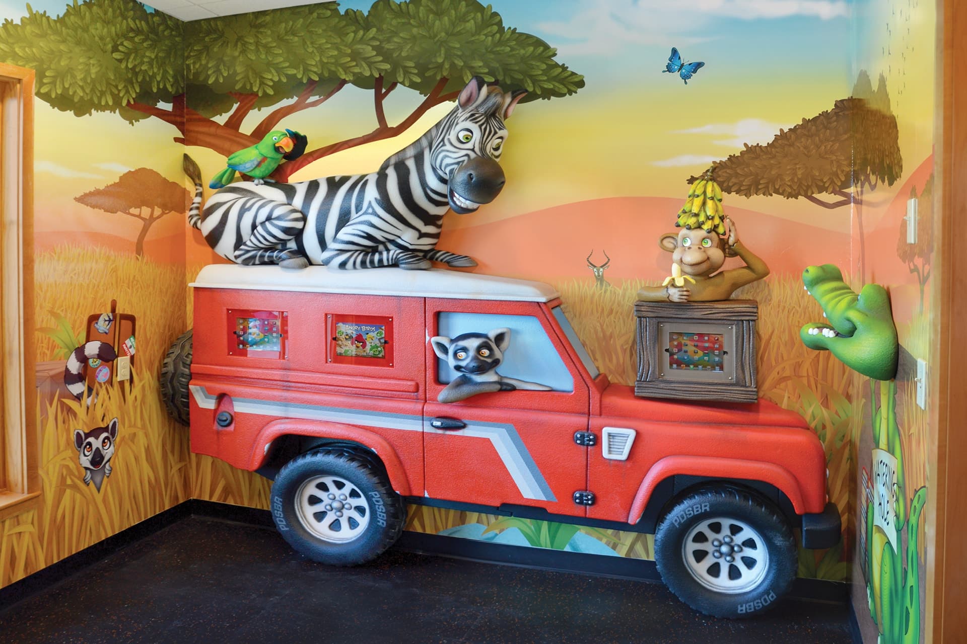 This vibrant kids space showcases a dramatic transformation—a red jeep built into the wall with a zebra, parrot, and monkey on top, a lemur inside, and a crocodile nearby, all set against lively savanna landscape murals.