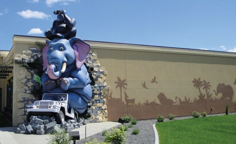 Large exterior landmark sculpture of an elephant driving a jeep with a gorilla seated on its head, bursting through the side of a building