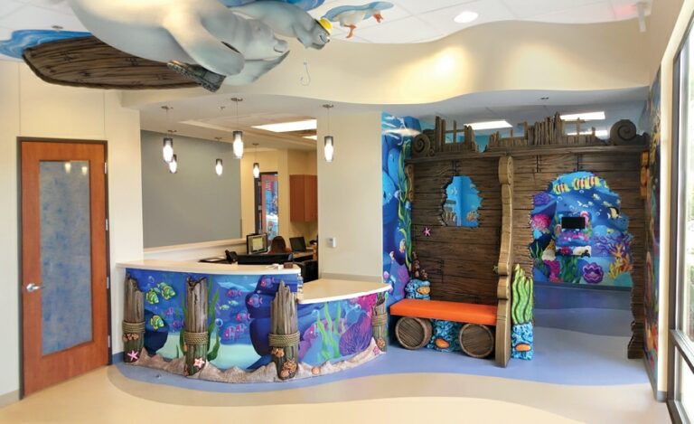 Ocean-themed reception/play area with underwater wall murals and sculpted shipwreck room divider. The desk has sculpted pier posts and beach sand cladding. A custom cushion seat with coral and wood barrel base sits off to the side and sculpted sea creatures are suspended from the ceiling.