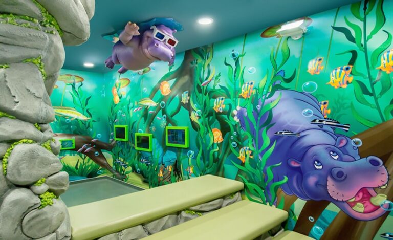 Jungle, underwater themed kids waiting area in a pediatric dental office. The decor includes wall murals, mounted gaming tablets, 3D sculpted rock benches and column with a ceiling-mounted hippo character swimming below the water line.