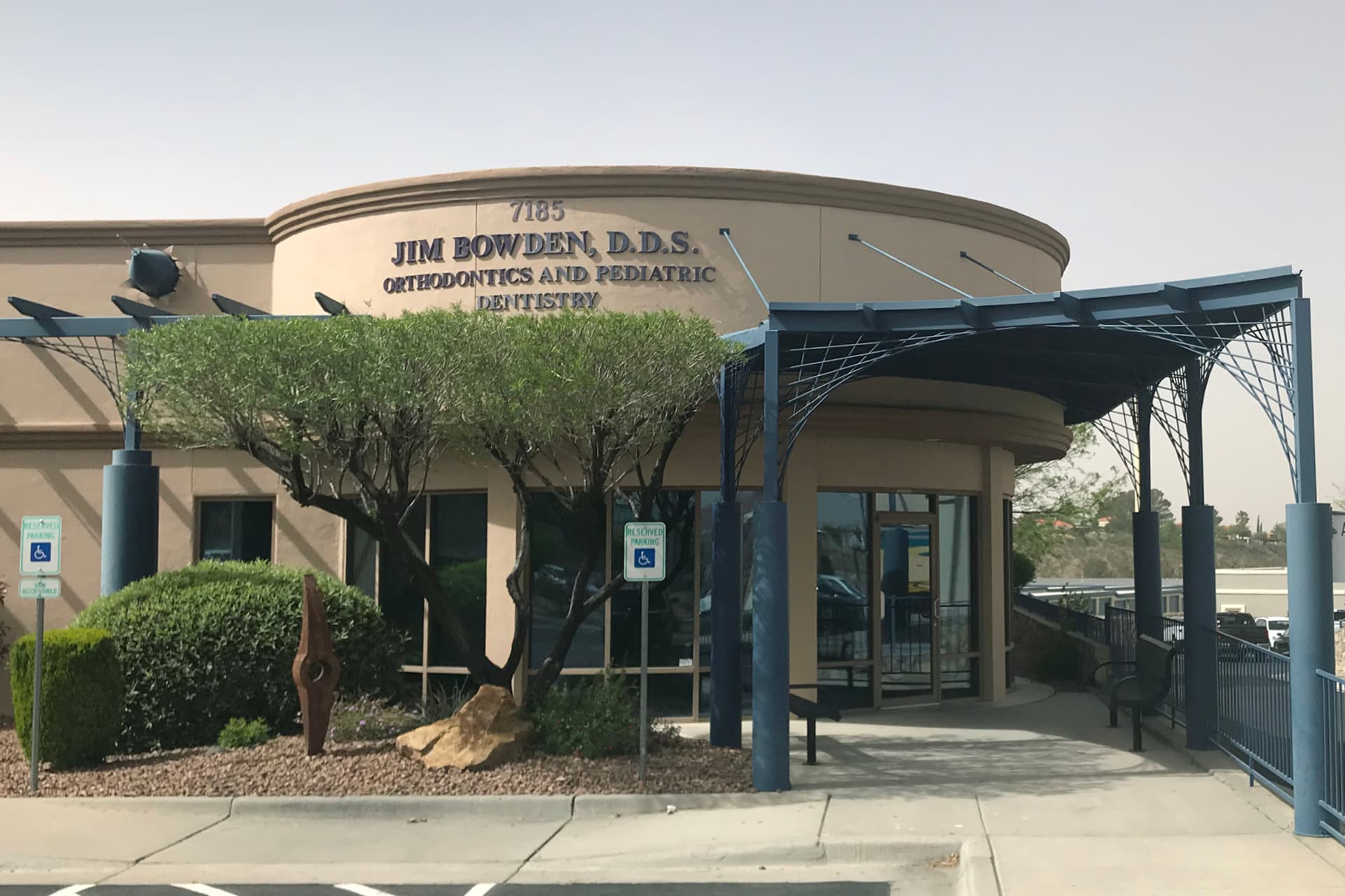 Before photo: The entrance of a dental office with a sign reading Jim Bowden, D.D.S. Orthodontics and Pediatric Dentistry appears drab, featuring trees, a metal sculpture, wheelchair ramp, and accessible parking spaces in front.