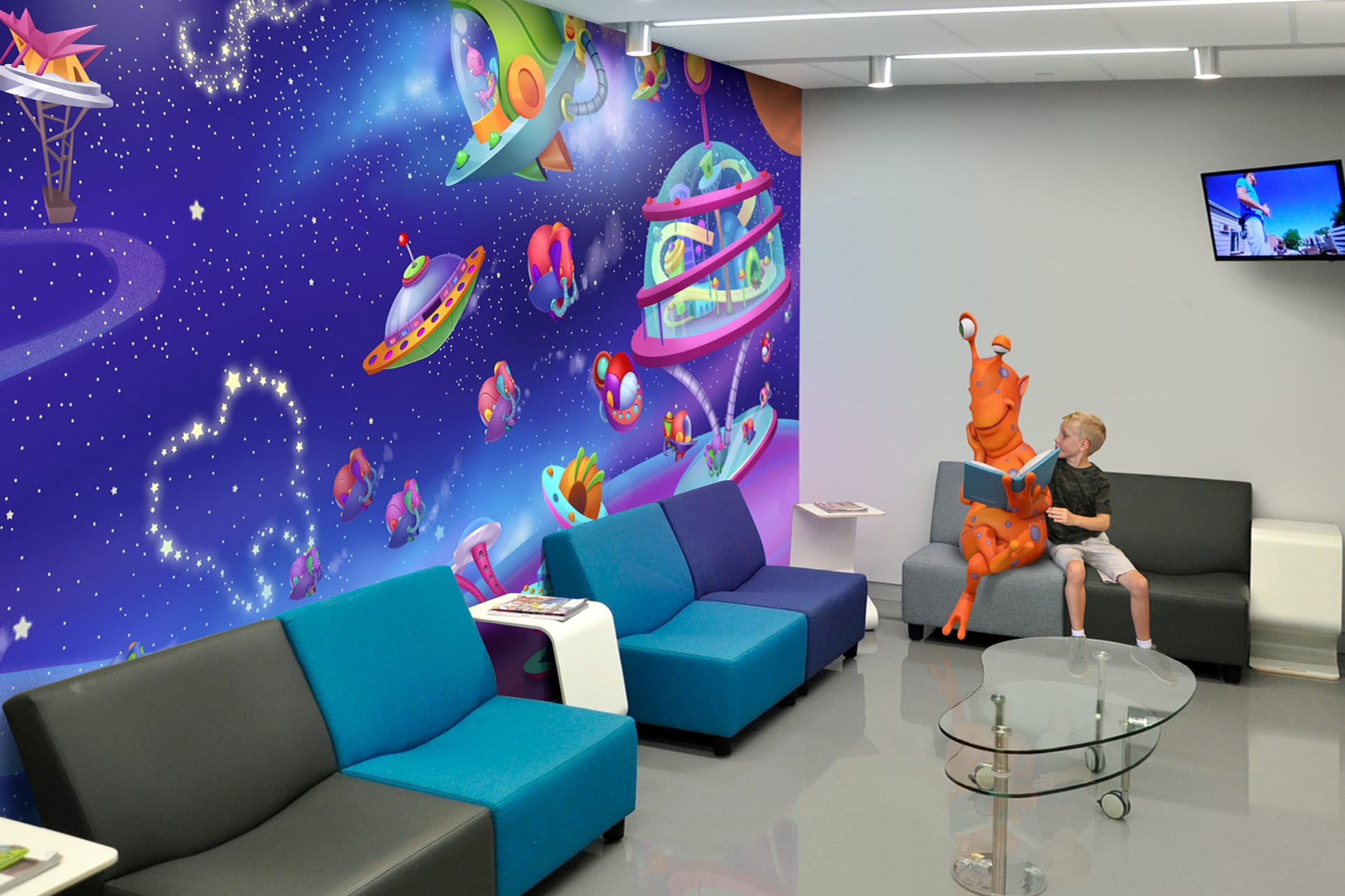 A colorful, space-themed mural with spaceships and aliens transforms this kids space, where blue and gray couches welcome visitors. In the after photo, a boy sits next to an orange alien statue, reading a book beneath a TV screen.