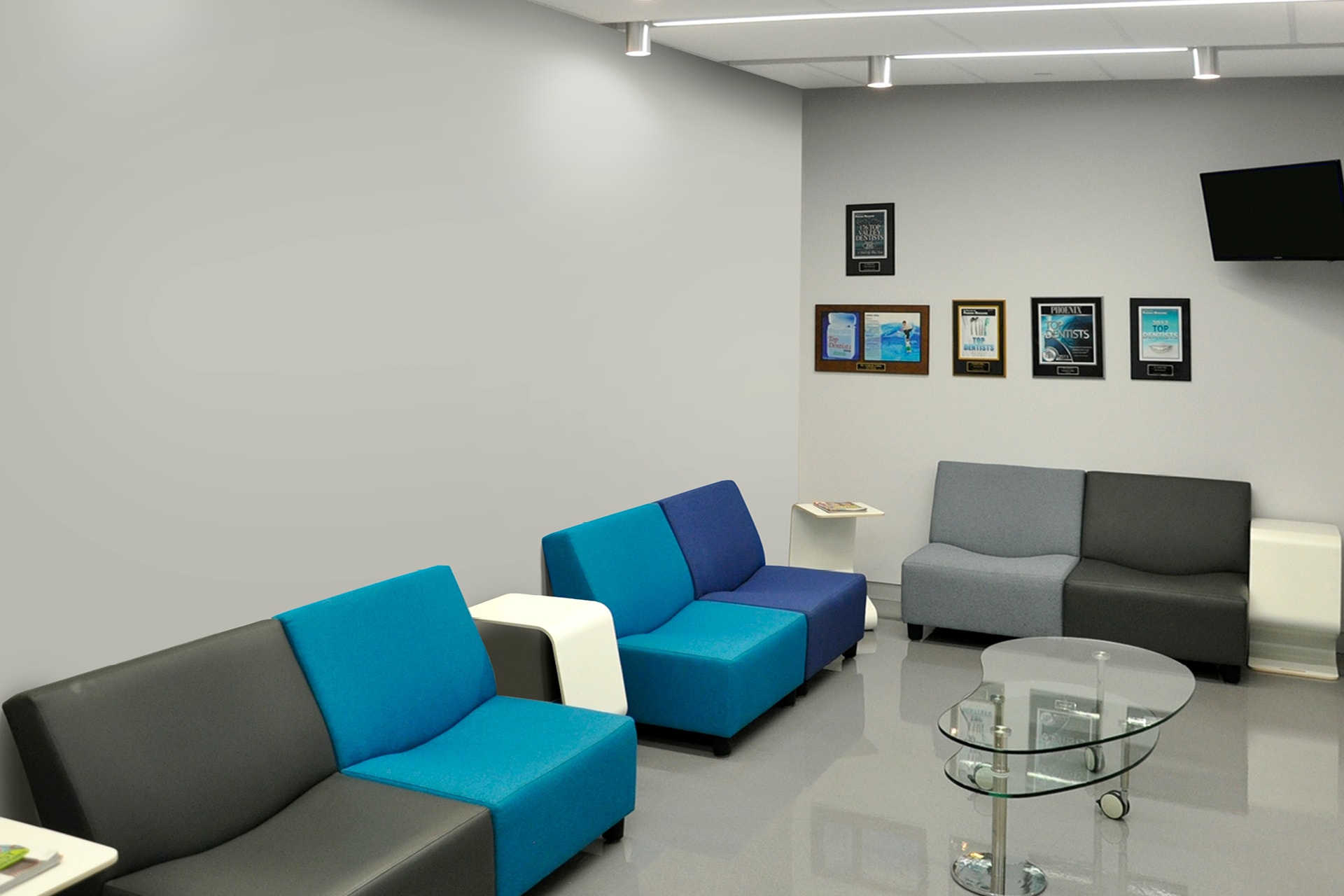 This before picture shows a modern waiting room with blue and gray sofas, a glass coffee table, wall plaques, and a wall-mounted TV. Despite its clean style, the space feels somewhat drab with neutral-colored walls and a polished floor.