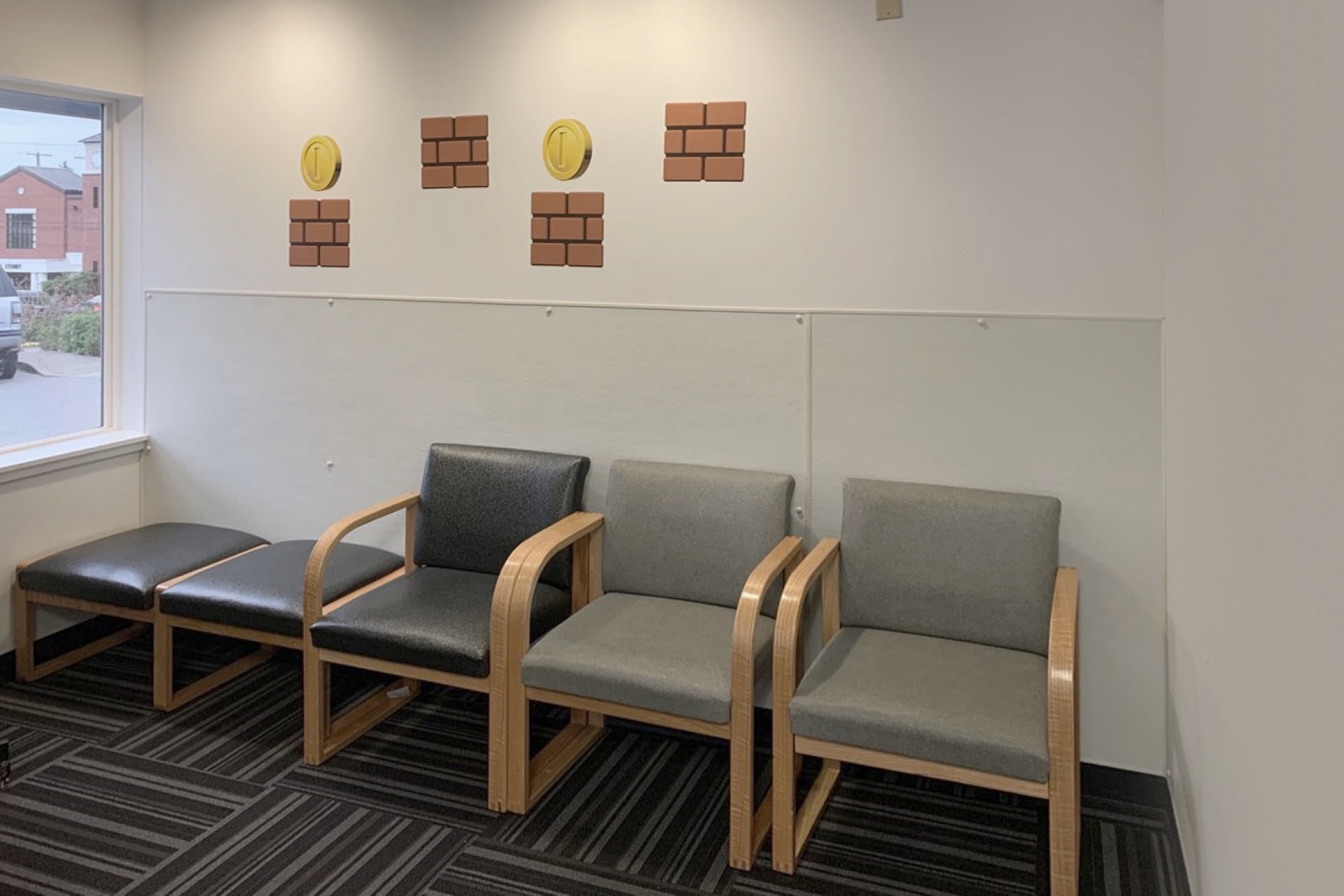 A drab waiting room with four gray chairs and one black chair against a white wall, decorated with Super Mario-style coin and brick decals. A window on the left lets in natural light—perfect for a