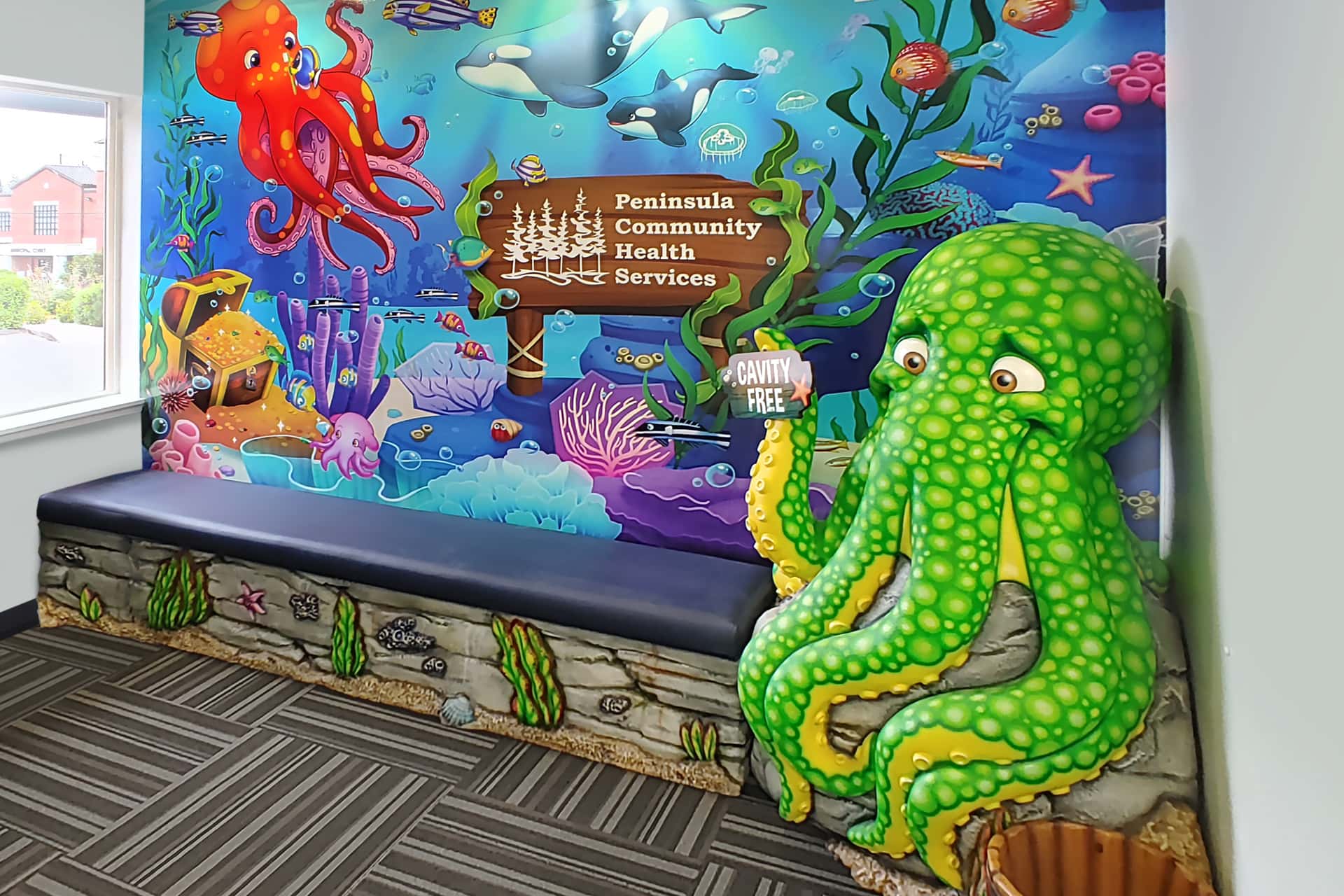 Colorful underwater-themed mural in a kids space waiting area with a large green octopus sculpture, fish, and sea creatures. A sign reads “Peninsula Community Health Services.” Cavity Free is on the octopus’s arm, showcasing the transformation.