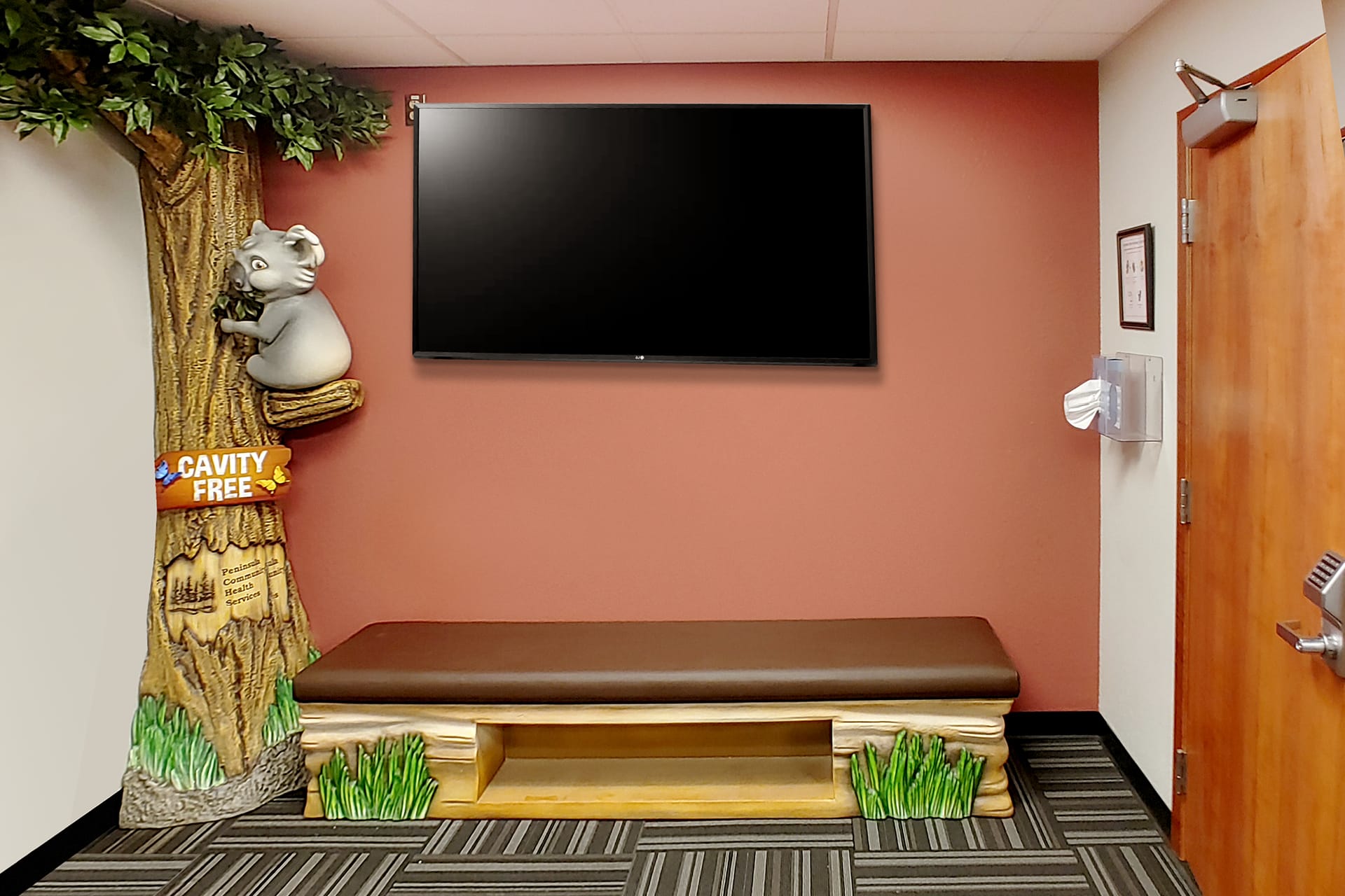 A waiting room featuring children’s decor—a bench shaped like a log, a tree with a koala and Cavity Free sign, a large TV on a red wall, and a door to the right shows the transformation in this inviting after photo.