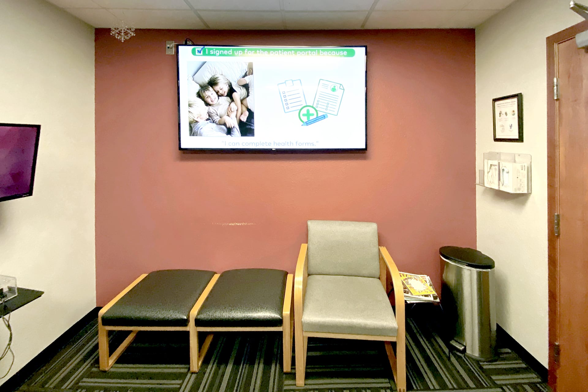 A small, drab waiting area with two cushioned benches, one armchair, a trash can, and magazines. Above the seating, a TV screen displays a sign-up message for the patient portal with images and forms; truly a “before photo.”.