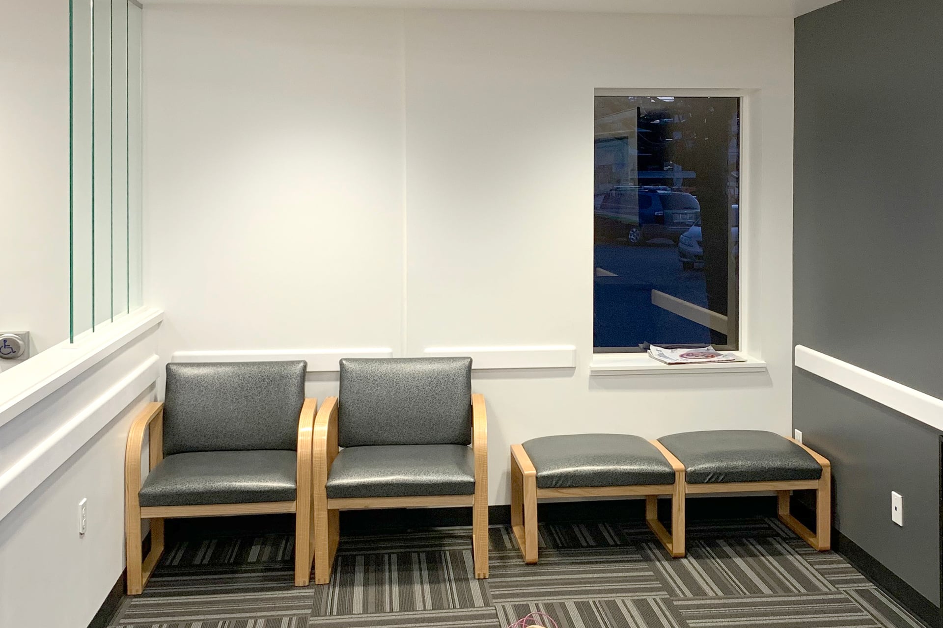 A drab waiting room with two gray cushioned chairs with wooden arms and matching benches. A window on the right holds a newspaper on the sill. White and gray walls and patterned carpet add to this boring before photo.