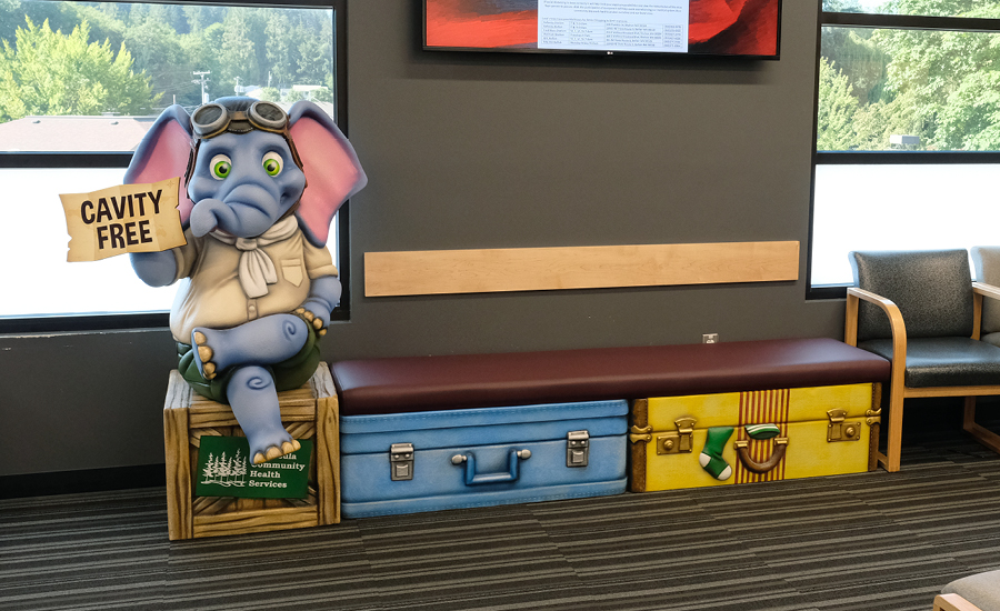 A whimsical elephant statue with goggles sits on a bench designed like colorful stacked suitcases, holding a Cavity Free sign in the dental and medical pediatric health center's waiting area, creating sculpted photo op characters for unique seating.