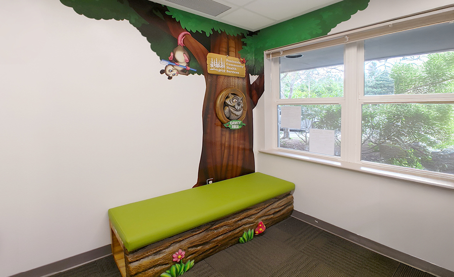 A children’s waiting area with a green bench designed like a log, a wall mural of a tree with a cartoon owl and squirrel, and a window with greenery visible outside.