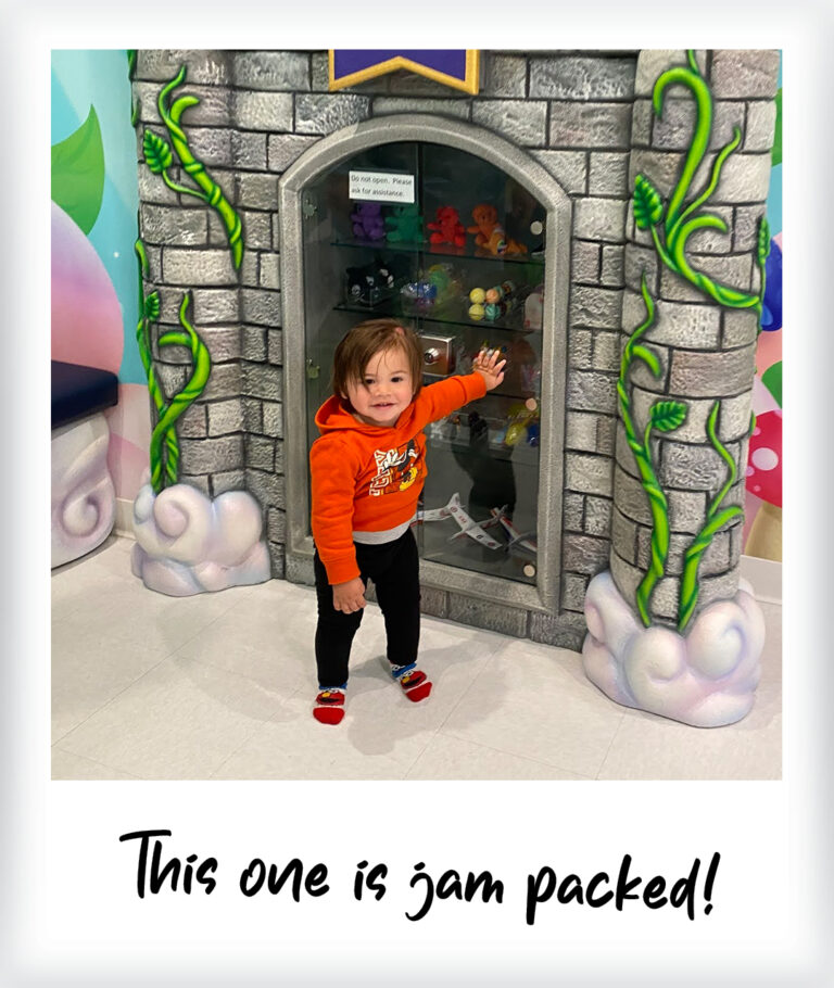 A smiling toddler in an orange hoodie points at a toy-filled display case shaped like a castle, set in vibrant play areas with painted vines and clouds. Text below reads, “This one is jam packed!”.