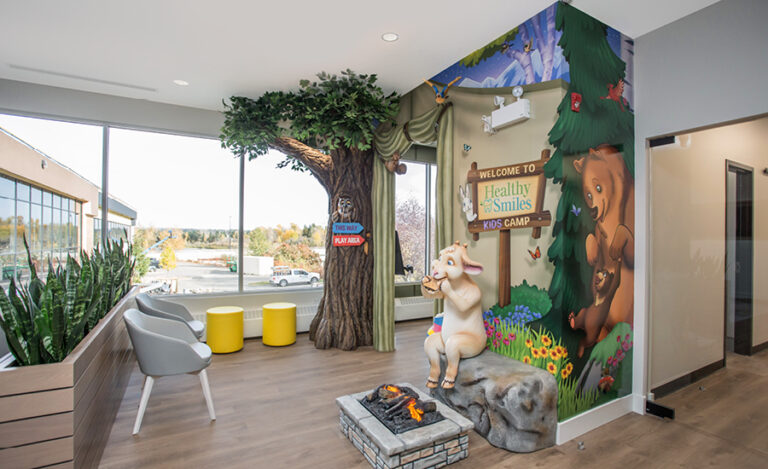 Cozy woodland themed entrance into a kid's play area with floor-to-ceiling sculpted tree and owl in the knothole. A sculpted goat character eats smores by a campfire. A colorful wall mural backdrop depicts a camp tent in the forest.