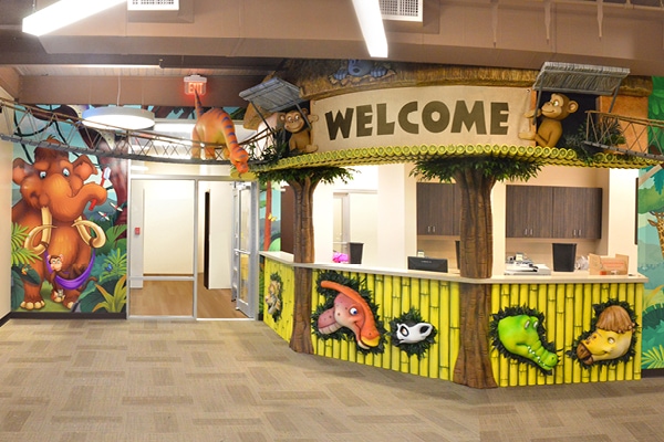 Dinosaur themed reception desk sided with yellow bamboo, 3D sculpted dinosaur heads, and a welcome sign.
