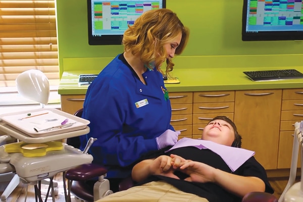 A happy dental staff member in blue scrubs speaks with a boy reclining in a dentist chair. The pediatric dental office features green cabinets, dental tools, and computer screens displaying colorful schedules in the background.