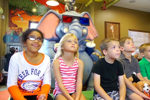 A group of children sit in front of a large cartoon elephant statue wearing 3D glasses, looking entertained and engaged at this vibrant jungle office. The background is colorful with playful, animated decor.