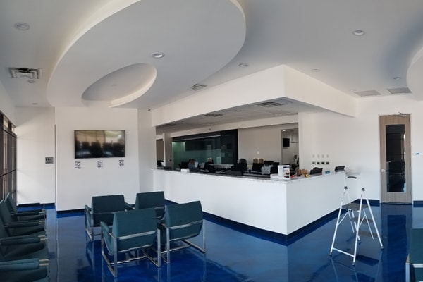 A drab, modern waiting area with blue flooring, several dark chairs, a white reception desk, wall-mounted TV, and contemporary ceiling design. A stepladder is placed near the door on the right in this before photo.