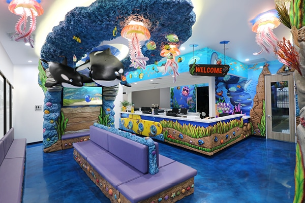 Colorful, underwater-themed waiting room with blue floors, purple benches, sea creature decorations, hanging jellyfish lights, and a WELCOME sign above a vibrant oceanic reception desk—an inspiring transformation in children’s decor.
