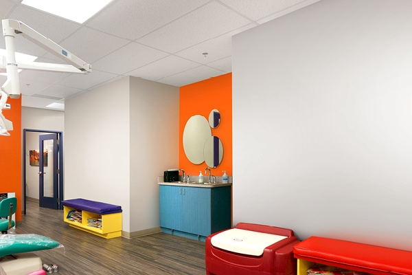 A brightly lit pediatric dental exam room with colorful furniture, an orange accent wall, round mirrors above a blue sink cabinet, and a hallway in the background; a definite upgrade from its previously drab and boring appearance in the before photo.