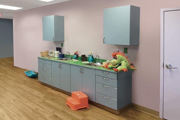 A drab children’s clinic exam room with light blue cabinets, a pink wall, two wall cabinets, toy animals on the counter, a step stool, a sink, and a wood floor—a perfect before photo.