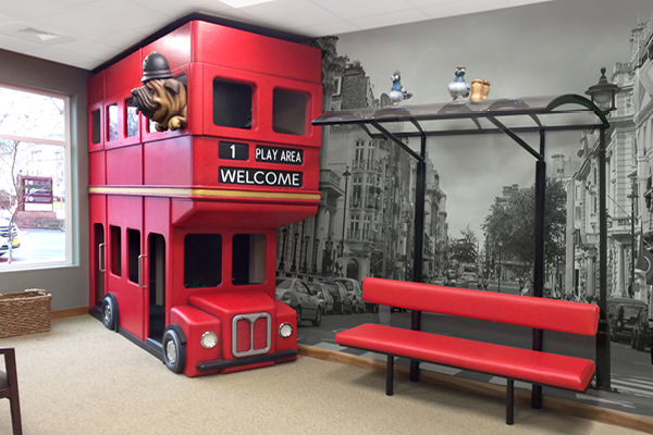 A children’s play structure shaped like a red double-decker bus sits beside a red bench under a faux bus stop canopy, all set against a city theme decor with a black and white mural of a bustling street.
