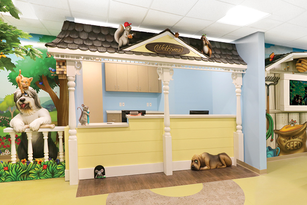 A whimsical, backyard-themed reception desk shaped like a small house with animal statues, including a dog, cat, and squirrel, decorates a brightly lit, playful childrens area with murals and colorful details.