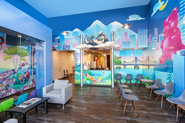 Colorful pediatric waiting room with a cartoon mural featuring mountains, animals, and a futuristic city. Reception desk under a cloud, small chairs and couches for all ages, bright blue walls, and playful decorations create a welcoming atmosphere.