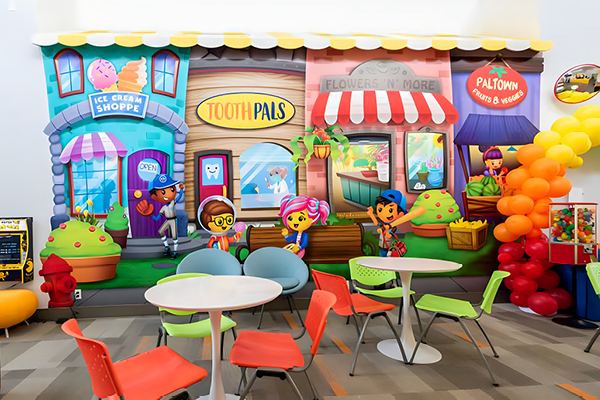 Colorful childrens dental office waiting area with town-themed decor and cartoon wall art of ice cream, dental, flower, and fruit shops. Bright chairs and tables fill the space, with a festive balloon arch on the right side.