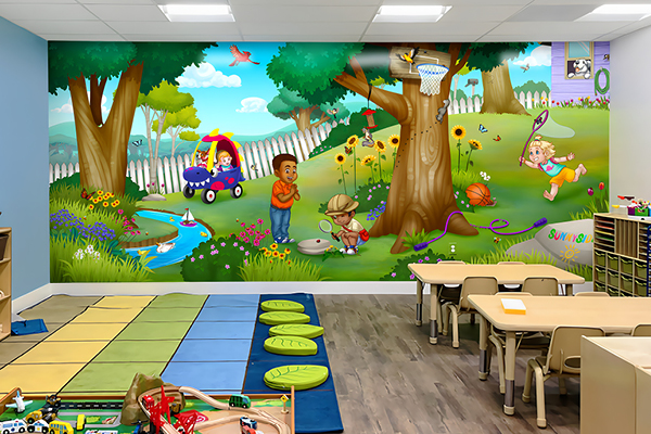 Colorful classroom with backyard-themed decor, featuring a large wall mural of children playing outside—using a wagon, playing basketball, chasing butterflies, and running. The room includes tables, chairs, mats, and toys.
