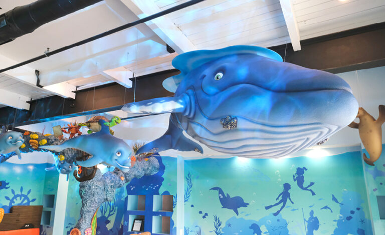 Large blue whale and sea creature sculptures hang from the ceiling in a brightly decorated undersea dental office waiting area, with ocean-themed murals and a striking blue silhouette mural on the walls.