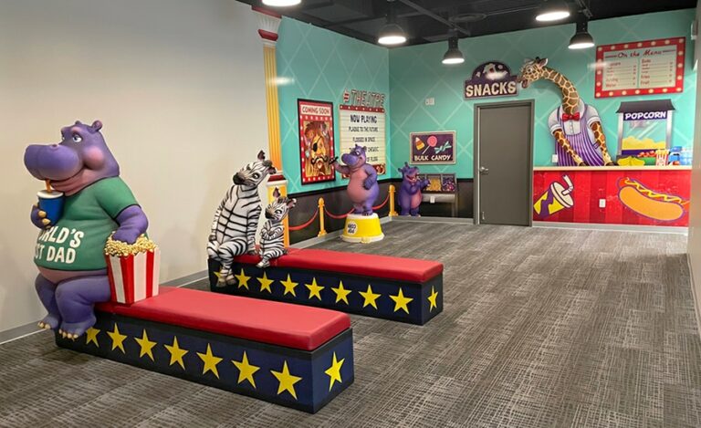 View of theater themed kid's waiting area with concession themed wall murals, sculpted hippo and zebra characters as well as custom bench seating.