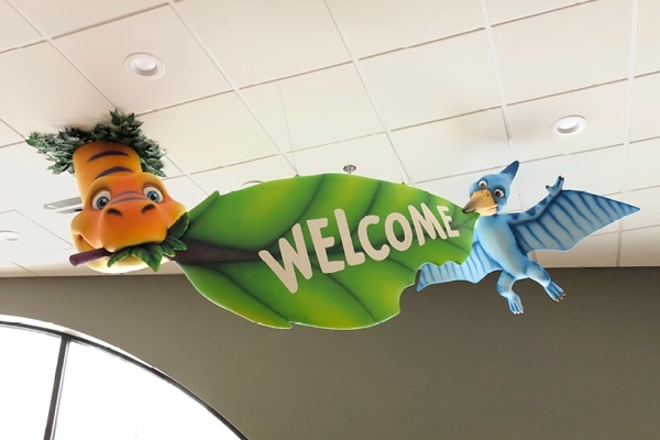 A leaf themed welcome sign being held up by a dinosaur pals.