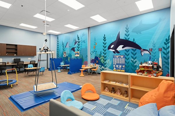 Colorful ABA therapy room for children with ocean-themed wall murals of orcas and fish, sensory swings, play mats, shelves with toys and books, a trampoline, and inviting activity stations—all brightly lit.