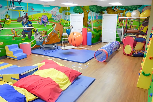 Colorful indoor pediatric physical therapy room with padded mats, bean bags, play tunnel, climbing wall, exercise balls, and swings. Cartoon animal murals decorate the walls, and there are various gym and sensory play equipment on a wood floor.