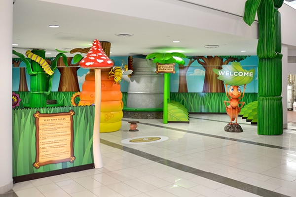 Indoor children’s play area with jungle-themed decorations, including large green leaves, a mushroom, a beehive, and a cartoon character holding a “Welcome” sign. This bright and inviting mall play area is perfect for young adventurers.