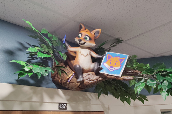 Door wayfinding sign. A cheerful fox statue sits on a branch above a doorway, holding a blue marker and a drawing of a fox face. Artificial green leaves surround the fox, and the ceiling is visible above.