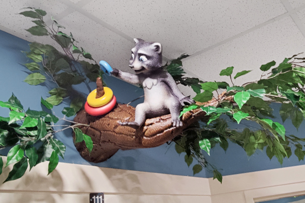 Door wayfinding sign. A sculpture of a raccoon sitting on a tree branch, reaching out to touch a blue ring stacked on top of red and yellow rings over a stick, with green leaves around them and a ceiling above.