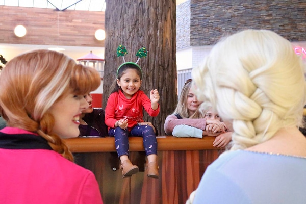 A young girl wearing holiday headbands smiles and waves excitedly at two women dressed as animated princesses in a mall play area, while adults stand nearby and watch the interaction.