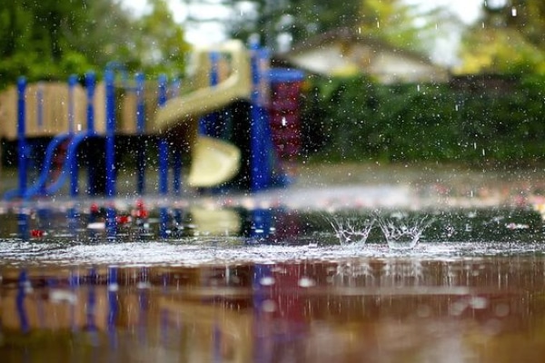 Raindrops splash on the wet ground in the foreground, with a blurred play area structure in the background, surrounded by green trees.