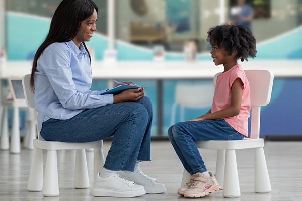 A woman holding a clipboard smiles and talks with a young girl in a speech therapy room. They sit on small white chairs facing each other indoors, both appearing engaged in conversation.