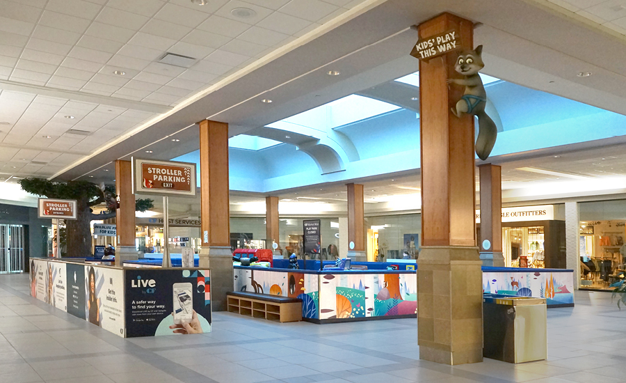 A vibrant kids zone in a shopping mall, bordered by colorful barriers and signs. A raccoon statue atop a column points to the business play area, while stroller parking and stores create a lively forest of activity in the background.