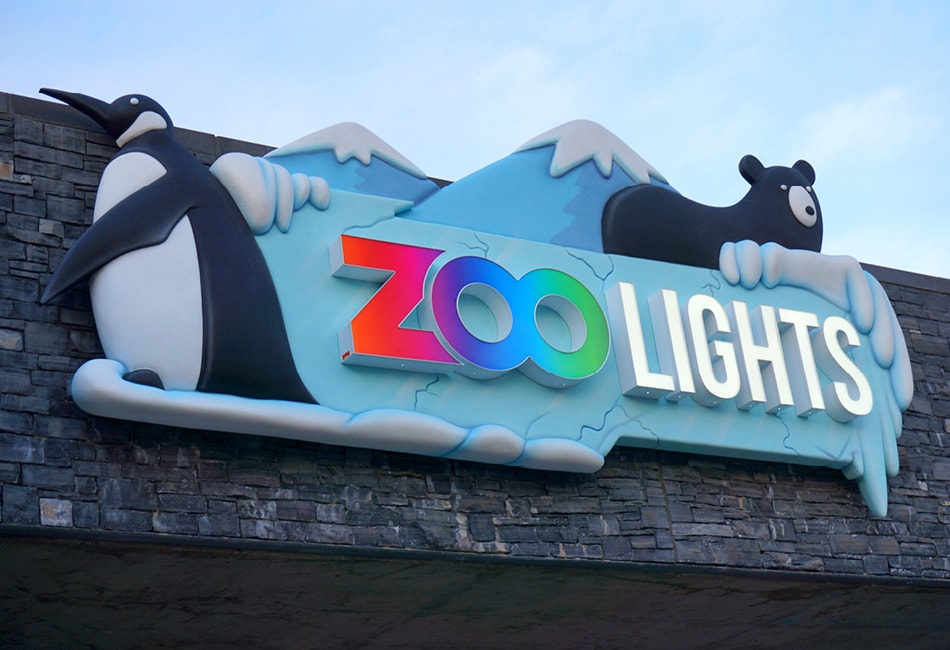 A colorful “Zoo Lights” sign features a penguin and a bear, with snowy mountains in the background, mounted on a stone wall—perfect for adding vibrant zoo decor to any family fun outing.